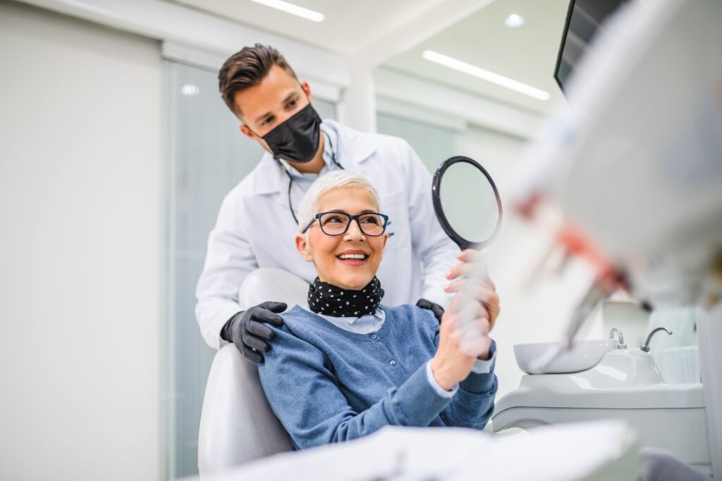 Woman smiling at reflection sitting in dental chair with dentist touching her shoulders